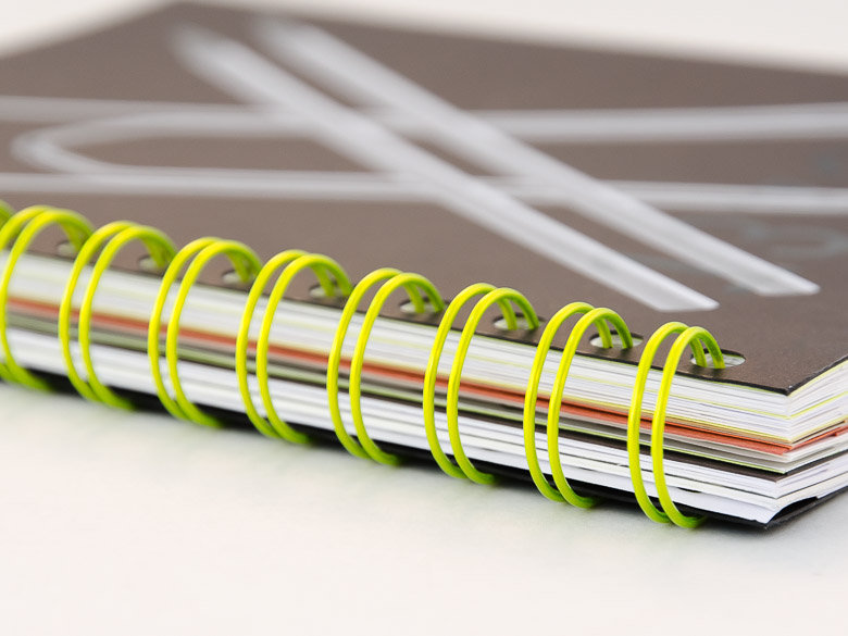 Wire-O binding with yellow coil Softcover notebook with bright green wire-o binding and multiple inner pages visible, lying flat on a white surface.