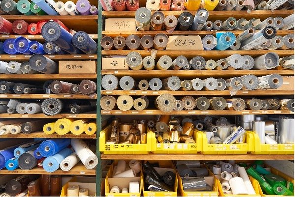 Regale mit zahlreichen Farbfäden auf Spulen in verschiedenen Farben und Materialien, darunter Blau, Gelb, Grau, Weiss und Braun, geordnet in Reihen und Fächern, darunter auch Rollen mit Folien oder Klebematerialien.