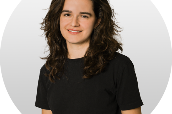 Person mit langen, lockigen dunklen Haaren trägt ein schwarzes T-Shirt und lächelt in die Kamera, vor einem hellgrauen, runden Hintergrund.
