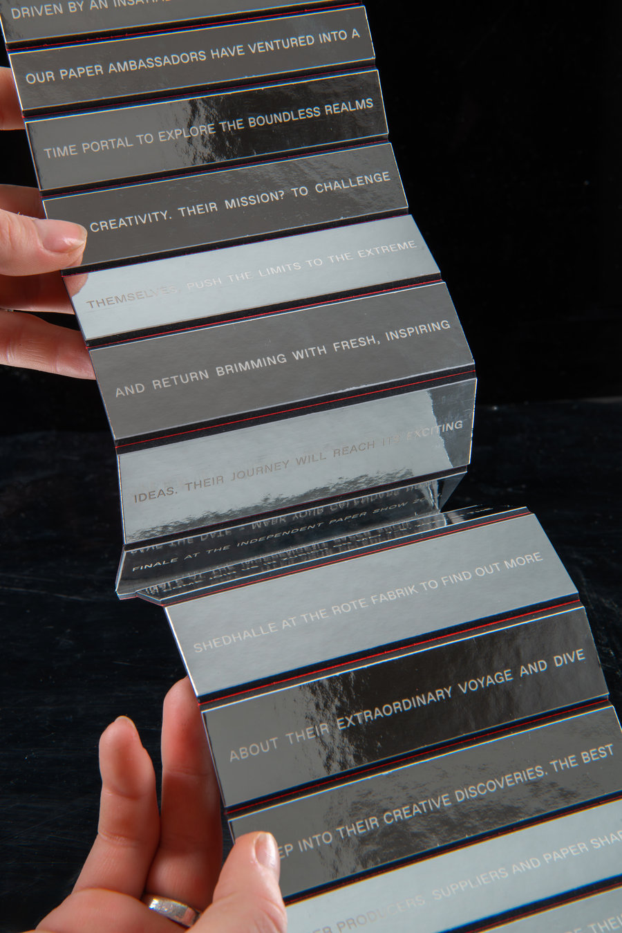 Livret accordéon transparent avec texte imprimé Deux mains tiennent un dépliant accordéon noir avec plusieurs volets contenant du texte blanc, chaque volet étant séparé par une fine ligne colorée rouge et bleue. Le fond est noir uni.