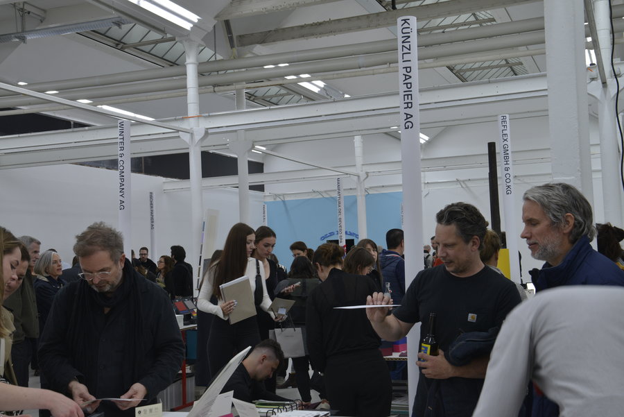 Salon professionnel de l'imprimerie et du papier Un grand espace intérieur avec plusieurs personnes debout autour de tables, examinant des documents, des livres ou des échantillons de papier. Des panneaux suspendus indiquent les noms de différentes entreprises liées au papier et à l’impression. Le décor comprend une structure métallique blanche et des murs clairs.