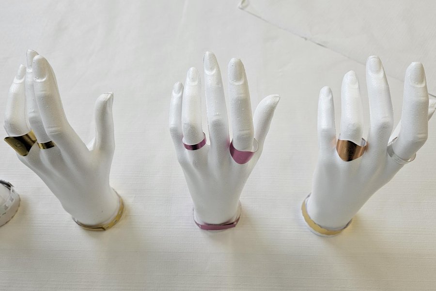 Présentoirs de mains pour bagues en vitrine Trois présentoirs de mains blanches en plastique portant chacun plusieurs bagues métalliques de différentes couleurs posés sur une surface claire.