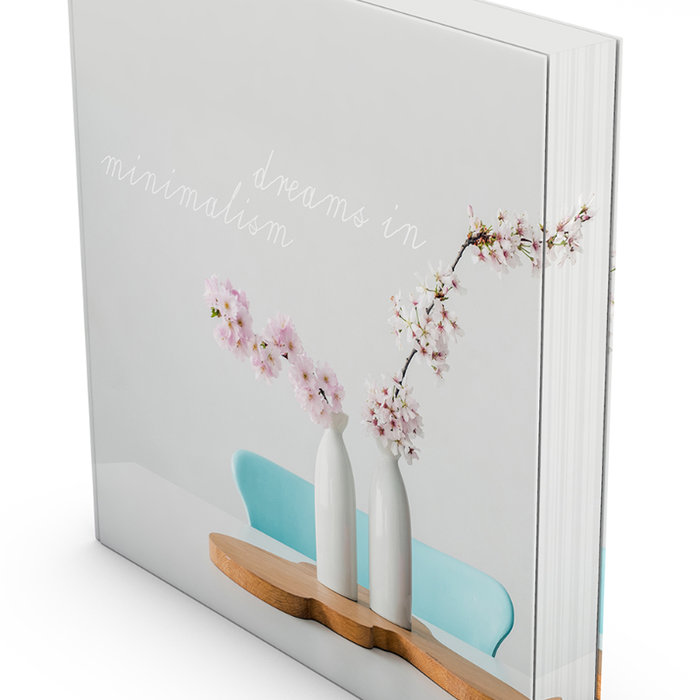 Quaderförmiger Softcover-Buchblock mit einem Coverfoto, das zwei weisse Vasen mit blühenden Kirschzweigen auf einem holzfarbenen Tisch vor einem hellblauen Stuhl zeigt. Auf dem Einband steht der Titel «dream me in minimalism» in einer feinen, weissen Schrift.