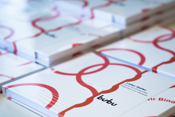 Softcover Bücherstapel mit Folienkaschierung Mehrere Hardcover-Bücher mit weissem Umschlag, darauf ein rotes geschwungenes Linienmuster und schwarzer Text mit dem Schriftzug "bubu". Die Bücher liegen stapelweise nebeneinander.