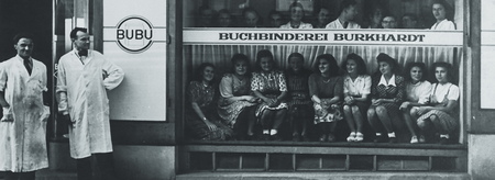 Schwarzes-Weiss-Foto vor dem Schaufenster der Buchbinderei Burkhardt mit mehreren sitzenden Personen hinter der Scheibe und zwei Männern in Kitteln links neben dem Eingang. Auf der Scheibe steht «Buchbinderei Burkhardt». Ein rundes Schild mit «BUBU» ist links zu sehen.