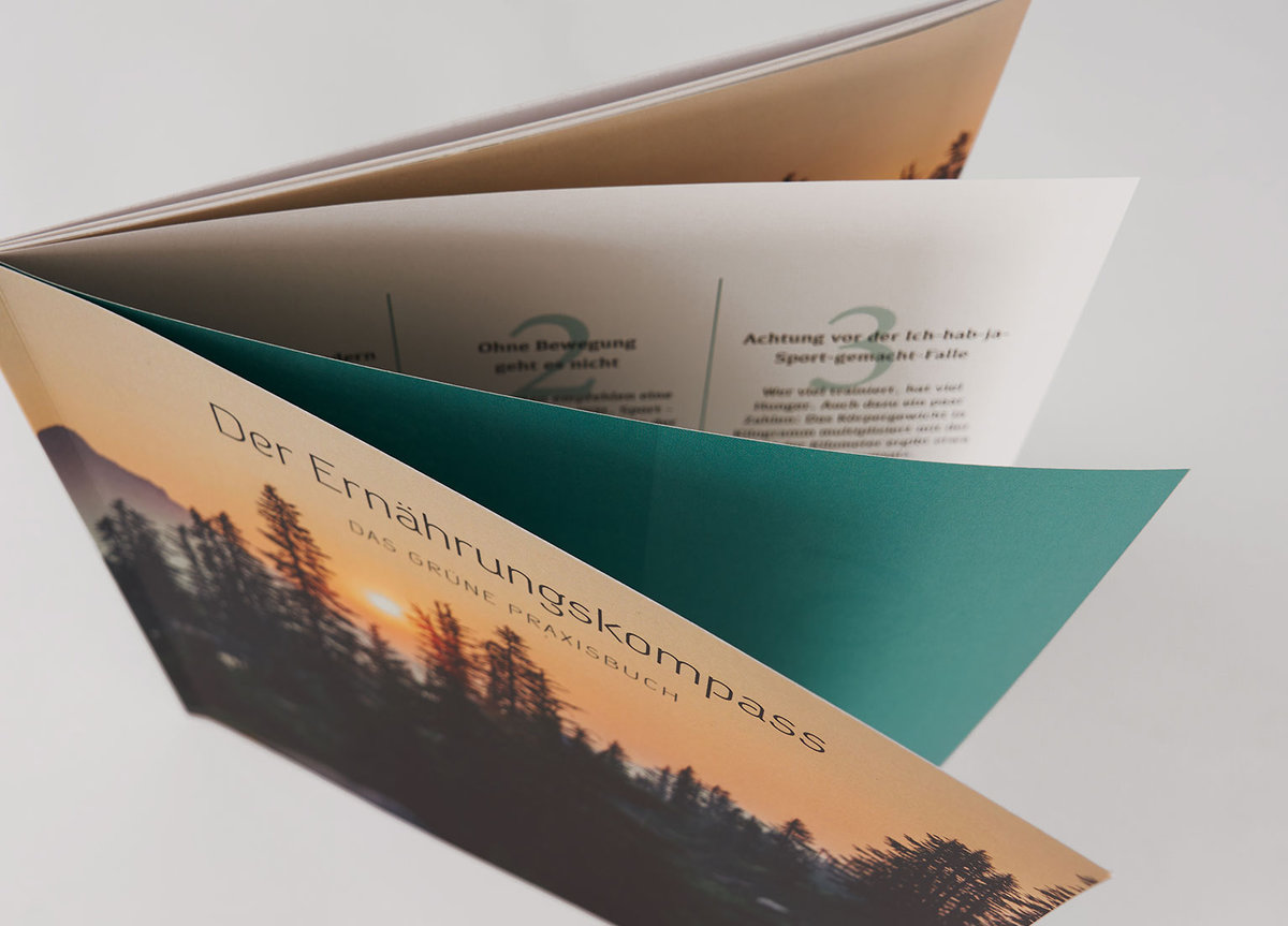 Dépliant plié en plusieurs volets avec une couverture illustrée d’un paysage forestier au coucher du soleil et le titre « Der Ernährungskompass ». Le dépliant montre du texte imprimé à l’intérieur sur fond blanc et un volet de couleur verte.