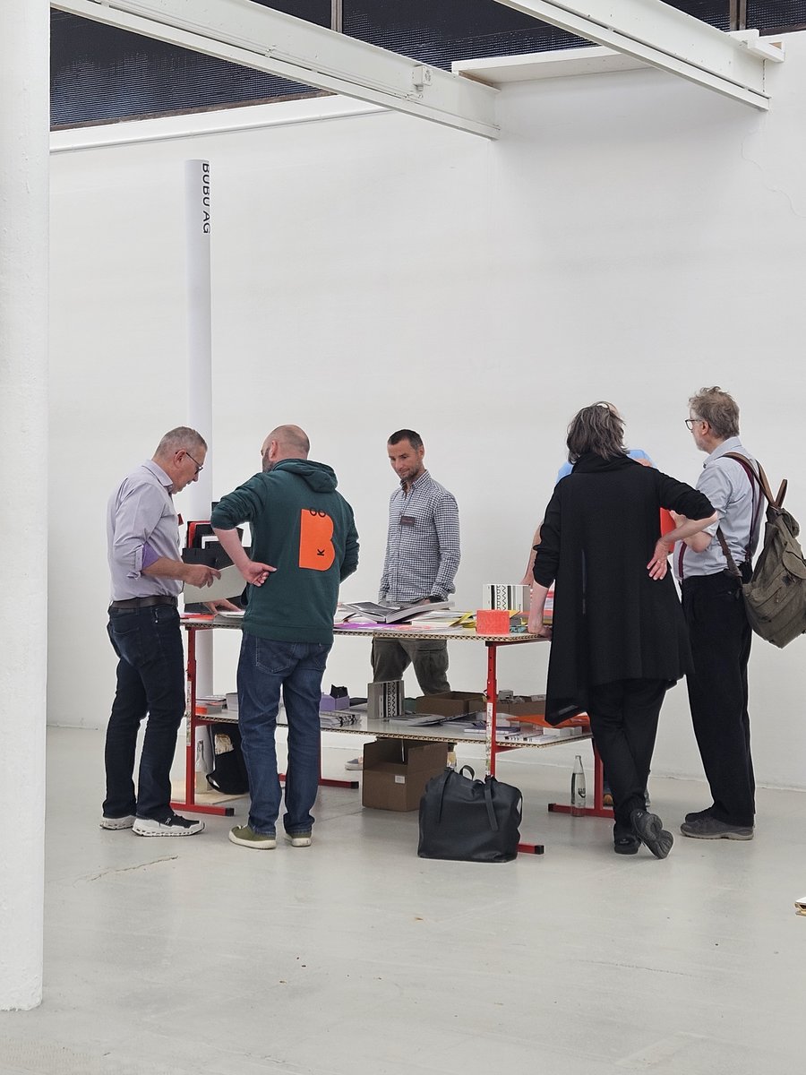 Rencontre autour de livres et documents Cinq personnes debout autour d'une table présentant des échantillons de matériaux et des ouvrages reliés, dans un grand espace blanc, probablement lors d'une exposition ou d'une réunion professionnelle.
