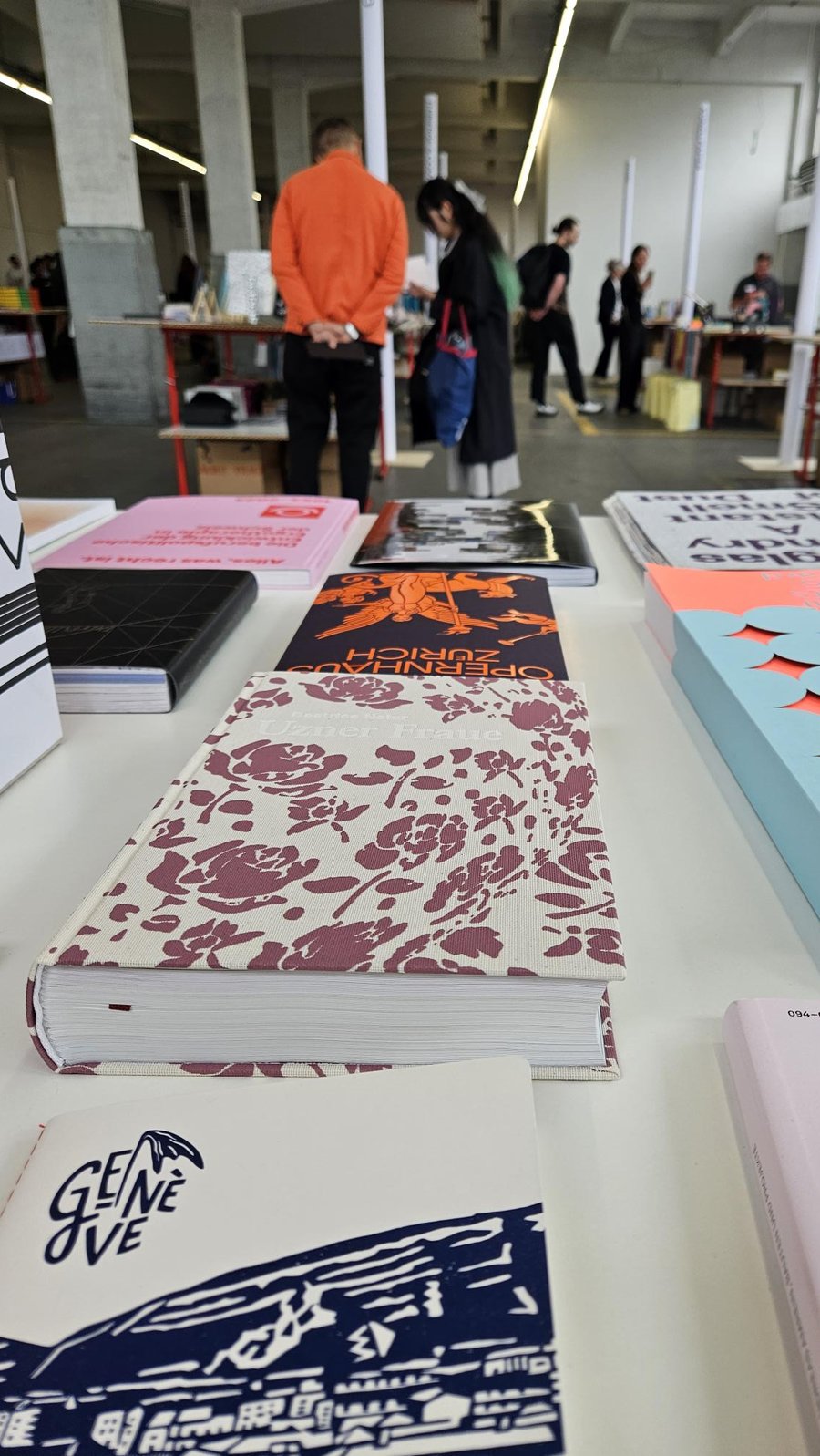 Livres exposés sur table blanche Plusieurs livres brochés et livres reliés sont posés sur une table blanche, présentant différentes couvertures colorées et motifs variés. En arrière-plan, plusieurs personnes se tiennent debout dans un espace intérieur lumineux avec des étagères et des matériaux d'impression visibles.