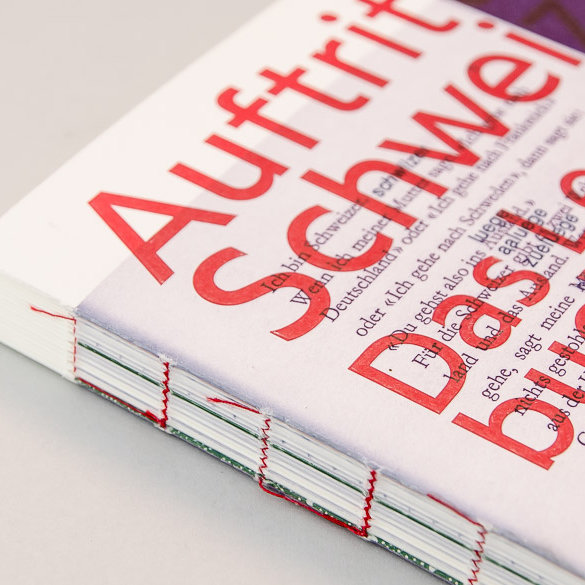 Fadenheftung mit rotem Faden an einem dickeren Softcoverbuch mit weiss-rotem Cover, der Buchrücken ist offen sichtbar.