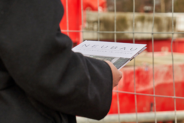 Brochure with title Neubau held by person Person holding a softcover brochure titled "NEUBAU" with additional text below the title, standing near a metal construction fence with red and grey construction site elements in the background.