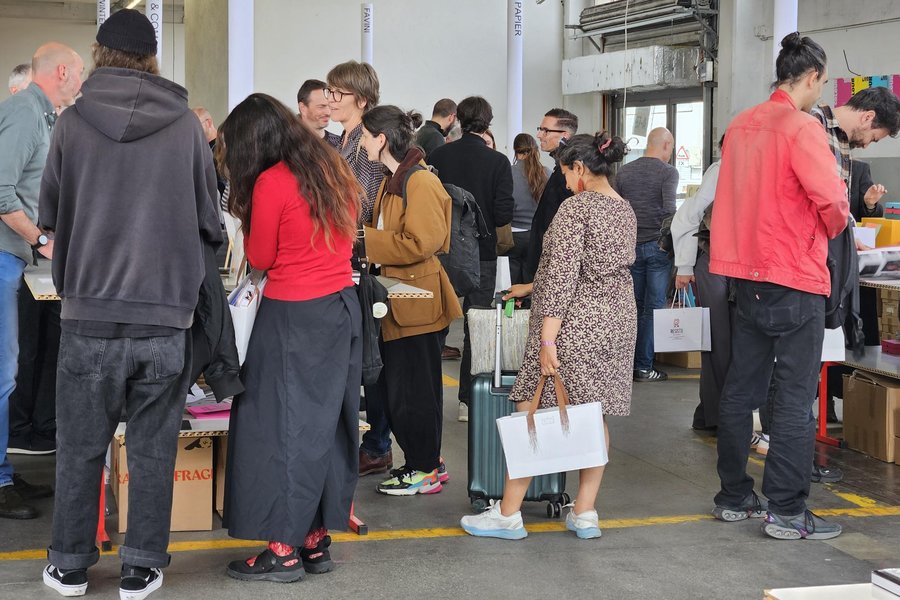 Personnes assistant à un salon professionnel en intérieur Plusieurs personnes se tiennent debout dans un grand espace intérieur, certaines tenant des sacs ou des feuilles, semblant en conversation ou en attente, dans un environnement qui pourrait être un atelier ou un hall de production.