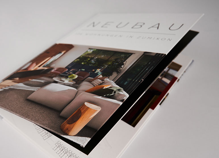 Folded brochure with interior design photos Softcover brochure with a photograph of a living room interior on the cover, showing a sofa, wooden side table, and large window with plants. The title "NEUBAU" is printed at the top of the front cover.