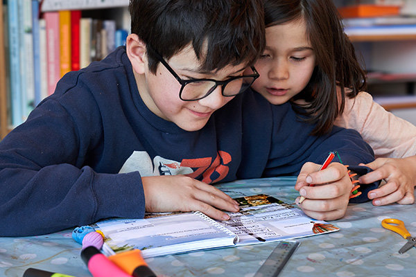 Enfants en train d'écrire et dessiner Deux enfants écrivent ensemble à la table, l'un tient un stylo rouge tandis que l'autre regarde attentivement sur un cahier ouvert. Des feutres et une règle sont posés sur la table autour d'eux.