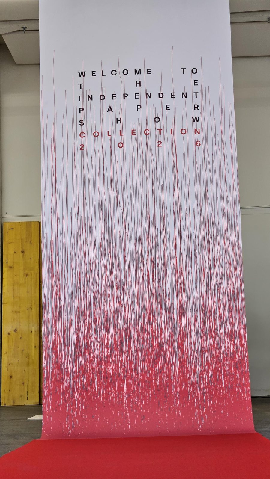 Affiche exposition papier indépendant 2026 Un grand rouleau de papier suspendu du plafond au sol, présentant un dégradé d’un rouge intense vers le bas à un blanc vers le haut, avec un texte imprimé en lettres noires et rouges au centre.
