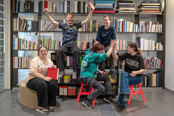 Fünf Personen sitzen und stehen vor einem grossen Regal mit vielen Büchern und gebundenen Produkten, einige halten Bücher in der Hand, zwei geben sich die Hände, eine Person freut sich mit erhobenen Armen.