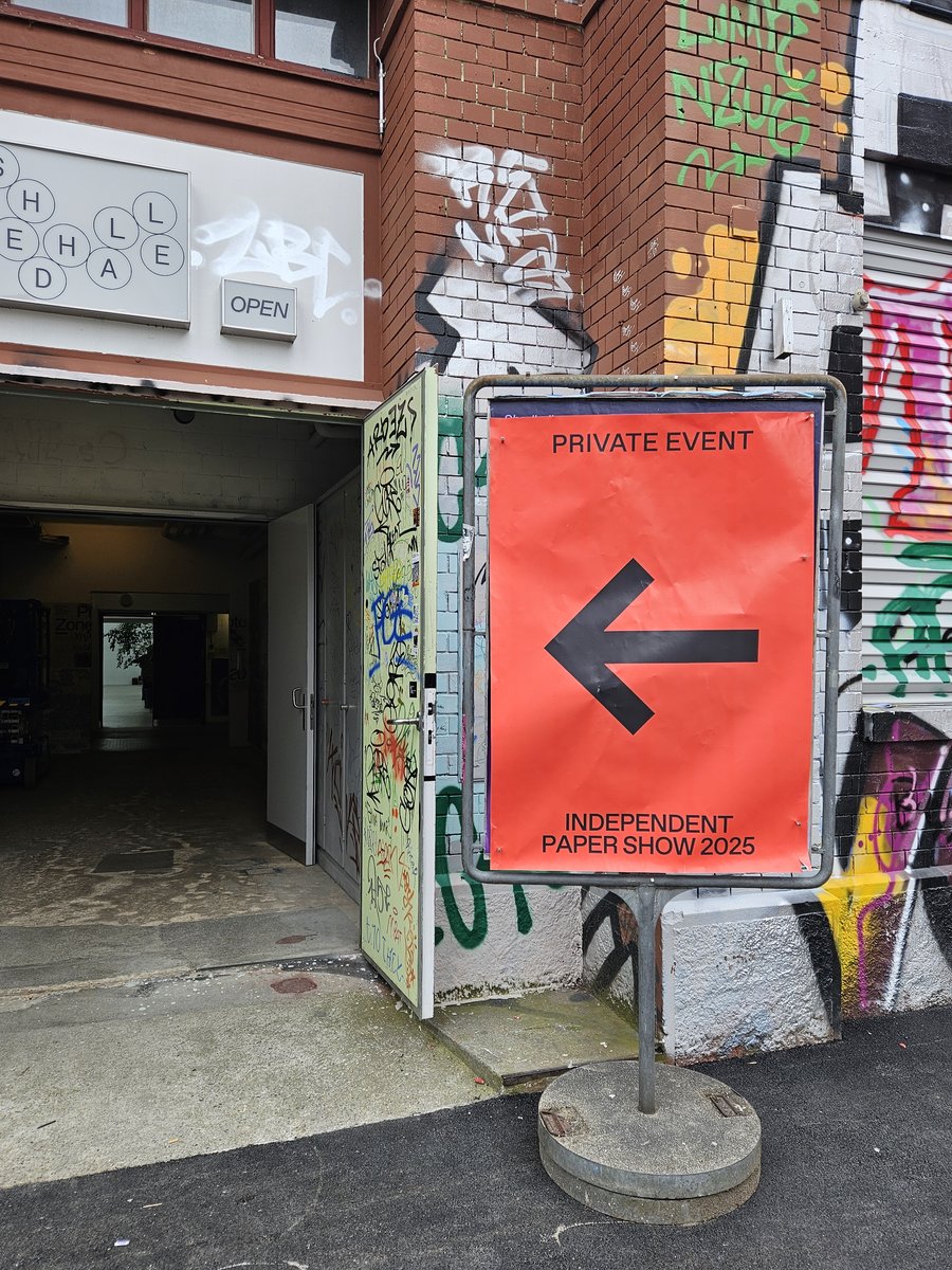 Signalisation extérieure pour exposition papier Affiche rouge indiquant un événement privé « Independent Paper Show 2025 » avec une flèche noire pointant vers la gauche, placée devant une entrée de bâtiment décorée de graffitis colorés sur les murs en briques et en béton.