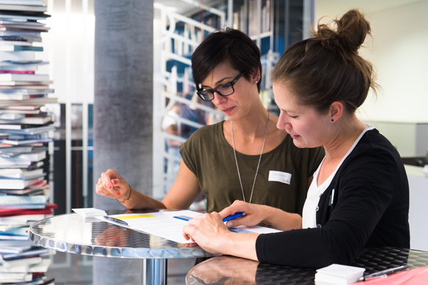 Teammeeting in der Buchbinderei Zwei Personen sitzen an einem runden Metalltisch und besprechen gemeinsam Dokumente. Im Hintergrund sind hohe Bücherstapel und Arbeitsbereiche erkennbar.
