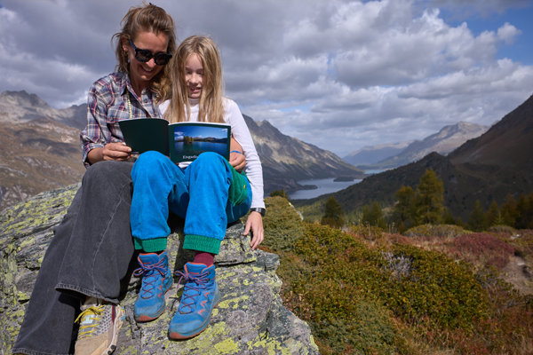 Lecture en plein air en montagne Deux personnes sont assises sur un rocher en pleine nature, l'une tient un livre ouvert qu'elles regardent ensemble, entourées de montagnes et d'un lac au loin sous un ciel nuageux.