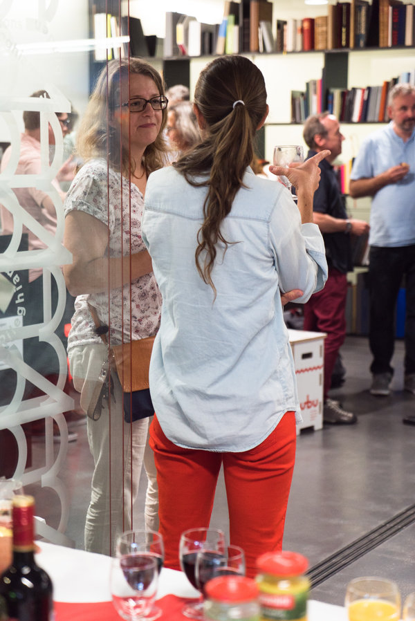 Gespräch bei Buchpräsentation Mehrere Personen unterhalten sich in einem Raum mit Regalen voll gebundener Bücher. Zwei Frauen im Vordergrund stehen dicht beieinander, eine hält ein Glas in der Hand. Auf einem Tisch sind Weinflaschen und Gläser zu sehen.