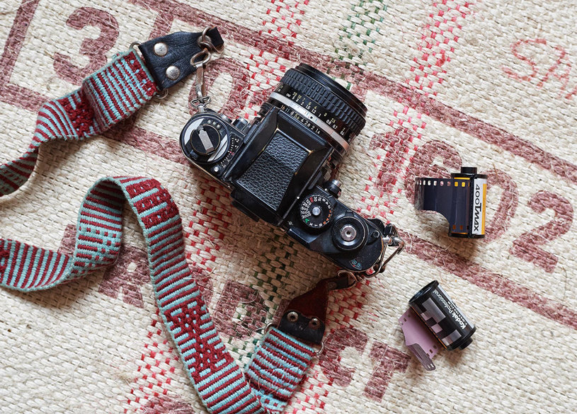 Analoge Kamera mit Filmrollen Eine analoge Spiegelreflexkamera mit Objektiv liegt auf einem gemusterten Stoff. Daneben liegen zwei Filmrollen, eine mit Film eingelegt und eine offene mit sichtbarem Negativstreifen. Die Kamera ist mit einem blau-roten Tragegurt ausgestattet.