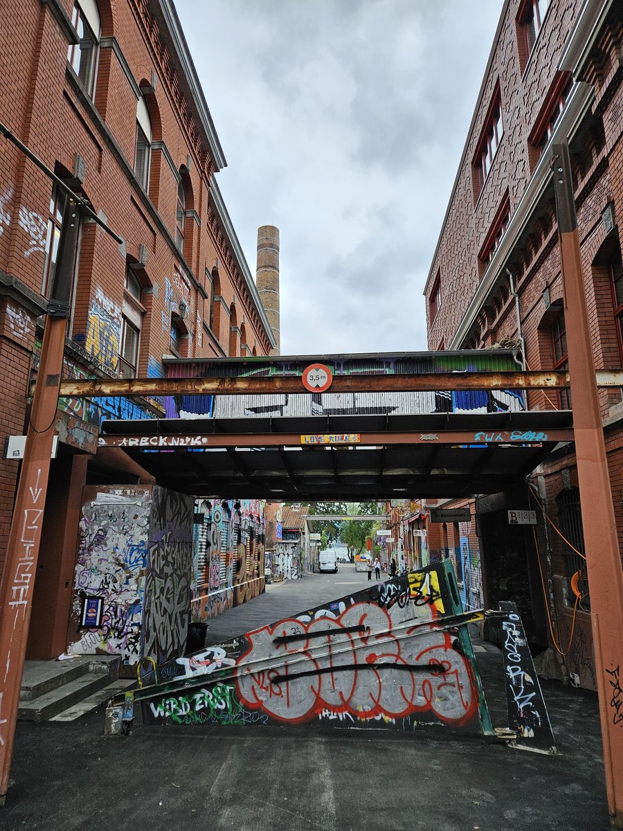 Passage urbain avec tag graffiti Allée étroite entourée de bâtiments en briques rouges avec des graffitis colorés sur les murs, barres métalliques et panneaux obstruant le passage au centre. Ciel nuageux visible au-dessus de la scène urbaine.
