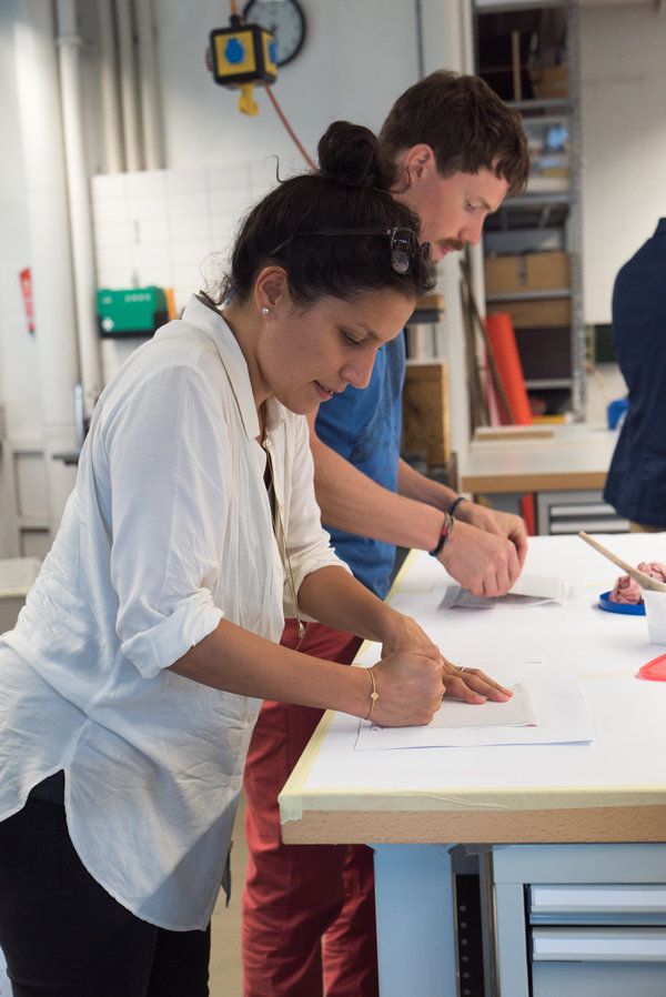 Menschen bei handwerklicher Arbeit am Tisch Zwei Personen stehen an einem Arbeitstisch und zeichnen mit Bleistift auf weisse Papierbögen, im Hintergrund Regale mit Materialien und Werkzeugen.