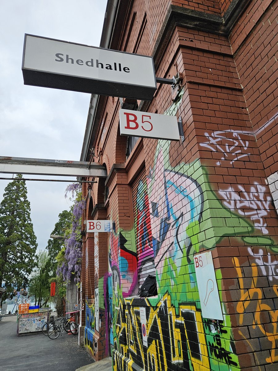 Façade en briques avec graffiti Bâtiment en briques avec plusieurs panneaux indiquant "Shedhalle", "B5" et "B6". Le mur est décoré de graffitis colorés. Des arbres, des vélos et un chemin sont visibles à côté du bâtiment.