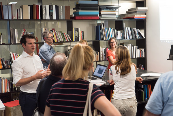 Meeting in Bücherregallager Eine Gruppe von Personen steht in einem Raum mit Regalen voller Bücher und Ordner. Zwei Personen diskutieren vor einem Laptop, andere hören zu. Der Raum wirkt wie ein Besprechungs- oder Arbeitsbereich in einer Buchbinderei oder Bibliothek.