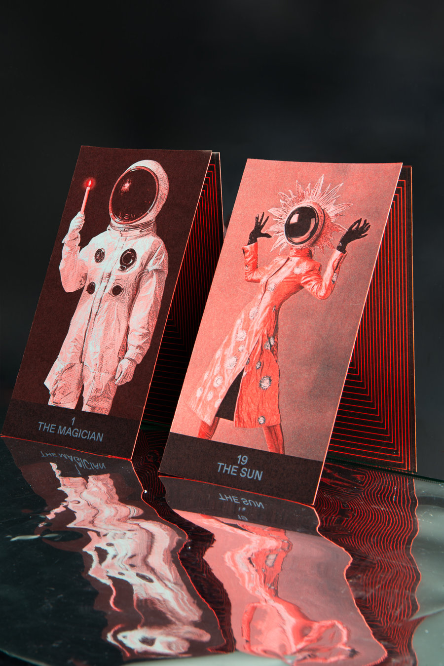 Cartes illustrées pliées debout Deux cartes illustrées debout, l'une intitulée "The Magician" montrant une personne en combinaison spatiale tenant un bâton lumineux, l'autre intitulée "The Sun" représentant une figure en robe avec un casque d'astronaute entouré de rayons lumineux. Les cartes sont posées sur une surface réfléchissante noire.