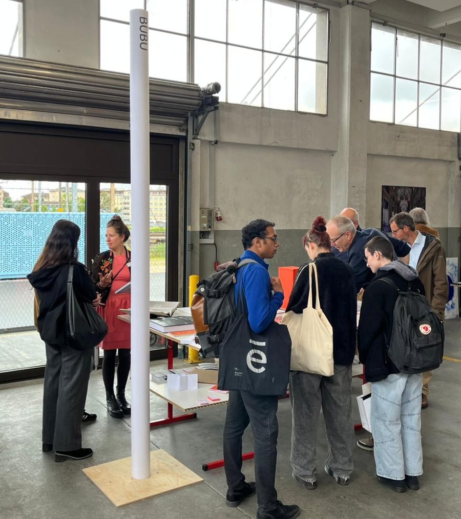 Événement de présentation en atelier Un groupe de sept personnes se tient autour d'une table dans un espace industriel lumineux avec de grandes fenêtres, semblant discuter ou examiner des documents ou objets posés sur la table. Un poteau blanc marqué "BUBU" est visible à proximité.