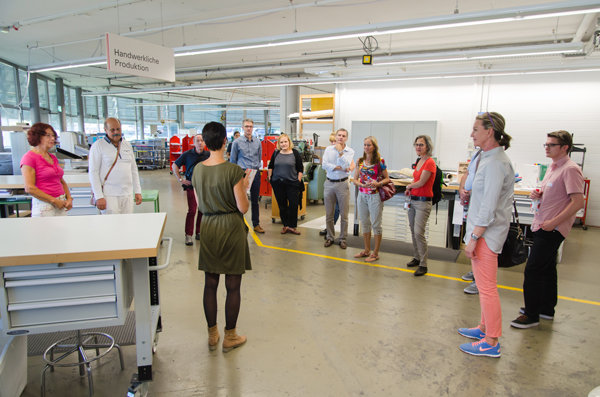 Führung durch handwerkliche Produktion Eine Gruppe von Personen steht in einer Werkhalle verteilt in einem Kreis und hört einer sprechenden Person zu. Im Hintergrund sind Arbeitsplätze, Regale und eine Tafel mit der Aufschrift „Handwerkliche Produktion“ zu sehen.