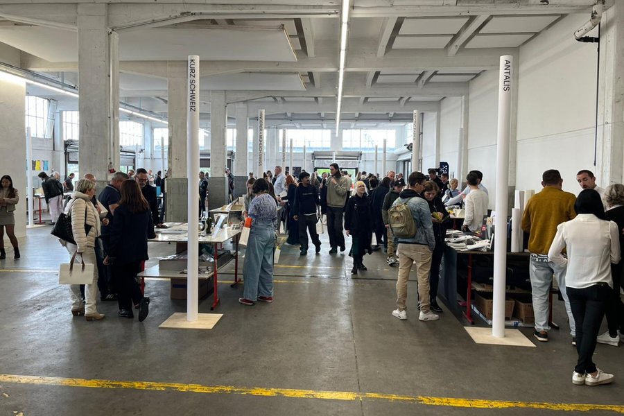 Salon professionnel dans un espace industriel Grand espace intérieur avec plusieurs groupes de personnes debout autour de tables présentant des objets ou documents, semblant discuter ou examiner des articles exposés. Deux panneaux blancs verticaux avec des noms de marques ou catégories sont visibles au centre.