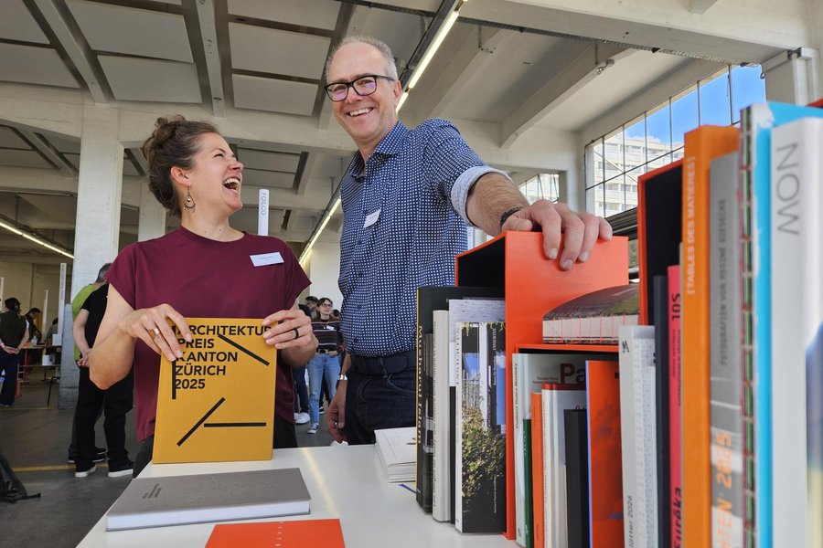 Présentation de livres avec couverture rigide colorée Deux personnes souriantes échangent autour d'une table où sont exposés plusieurs livres brochés et reliés, dont un exemplaire orange en couverture rigide tenant dans les mains de la femme à gauche.