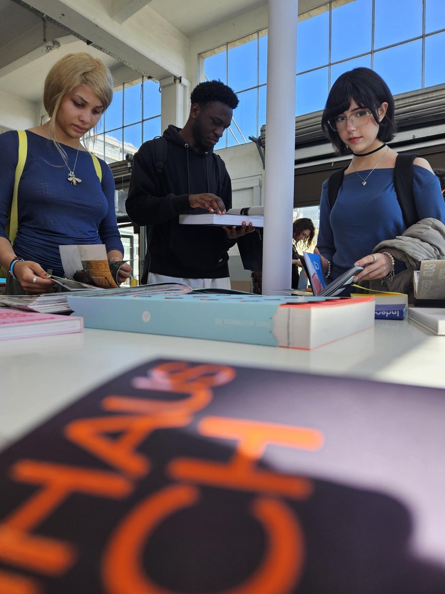 Personnes feuilletant des livres sur une table Trois personnes consultent des livres posés sur une table, dans un espace lumineux avec de grandes fenêtres. Plusieurs livres ouverts et fermés sont visibles sur la table blanche, au premier plan un livre avec une couverture sombre portant un titre en lettres orange.