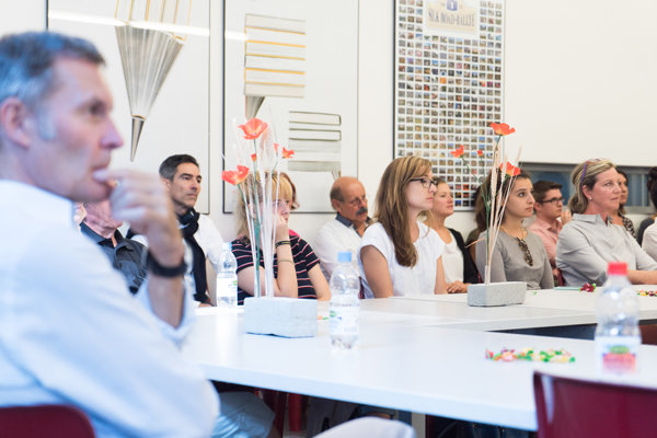 Personen im Seminarraum bei Vortrag Mehrere Personen sitzen an weissen Tischen in einem Raum und hören aufmerksam zu. Auf den Tischen stehen dekorative Vasen mit roten Kunstblumen sowie Wasserflaschen. Im Hintergrund sind an der Wand gerahmte Bilder und Poster zu sehen.