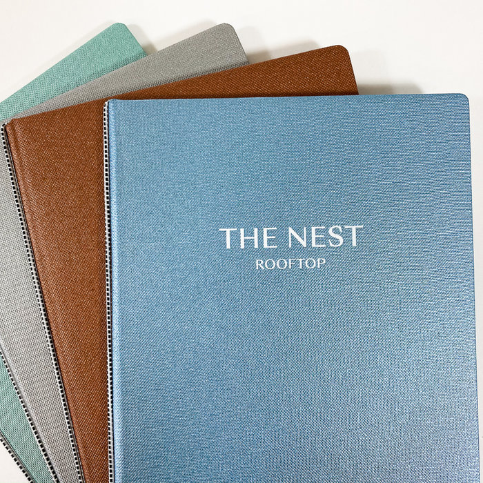 Vier Hardcover-Bücher mit Leineneinband in den Farben Blau, Braun, Grau und Grün, auf weissem Untergrund liegend, Titel "THE NEST ROOFTOP" auf dem blauen Buch in weisser Prägung.