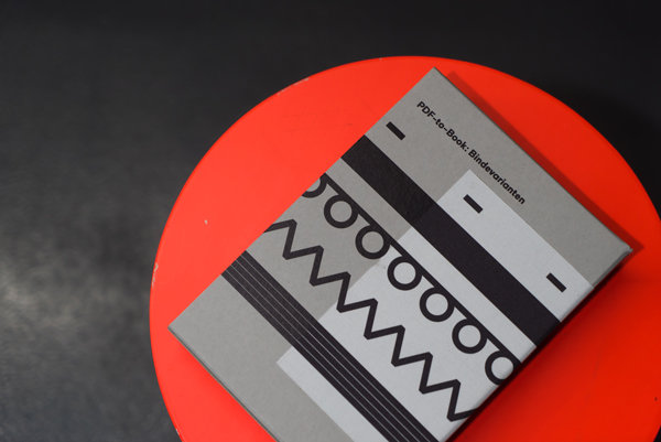 Softcover Buch mit grafischem Einband Ein Buch mit einem grauen Cover, das verschiedene Bindungsvarianten in Schwarz zeigt, liegt auf einem roten runden Untergrund vor schwarzem Hintergrund.